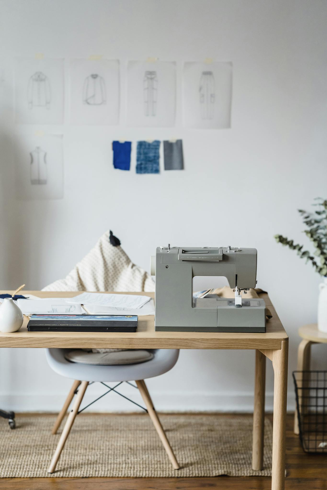 Professional sewing machine on wooden table near chair at wall with drafts of clothes and samples of textile in atelier